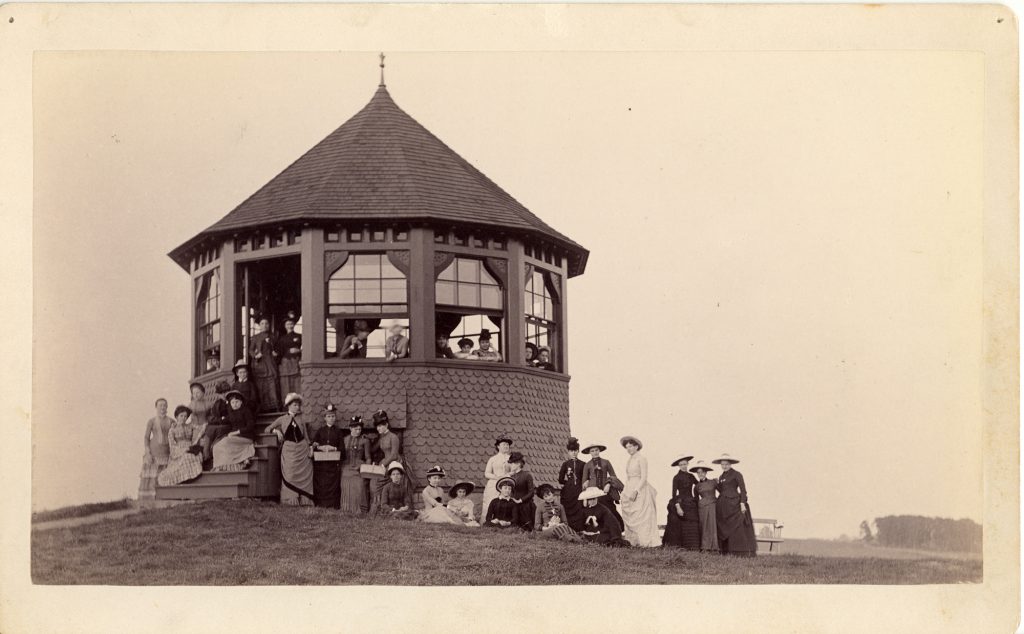 Women dressed in dresses and hats gather around an octagonal structure with a conical roof, posing for the picture.