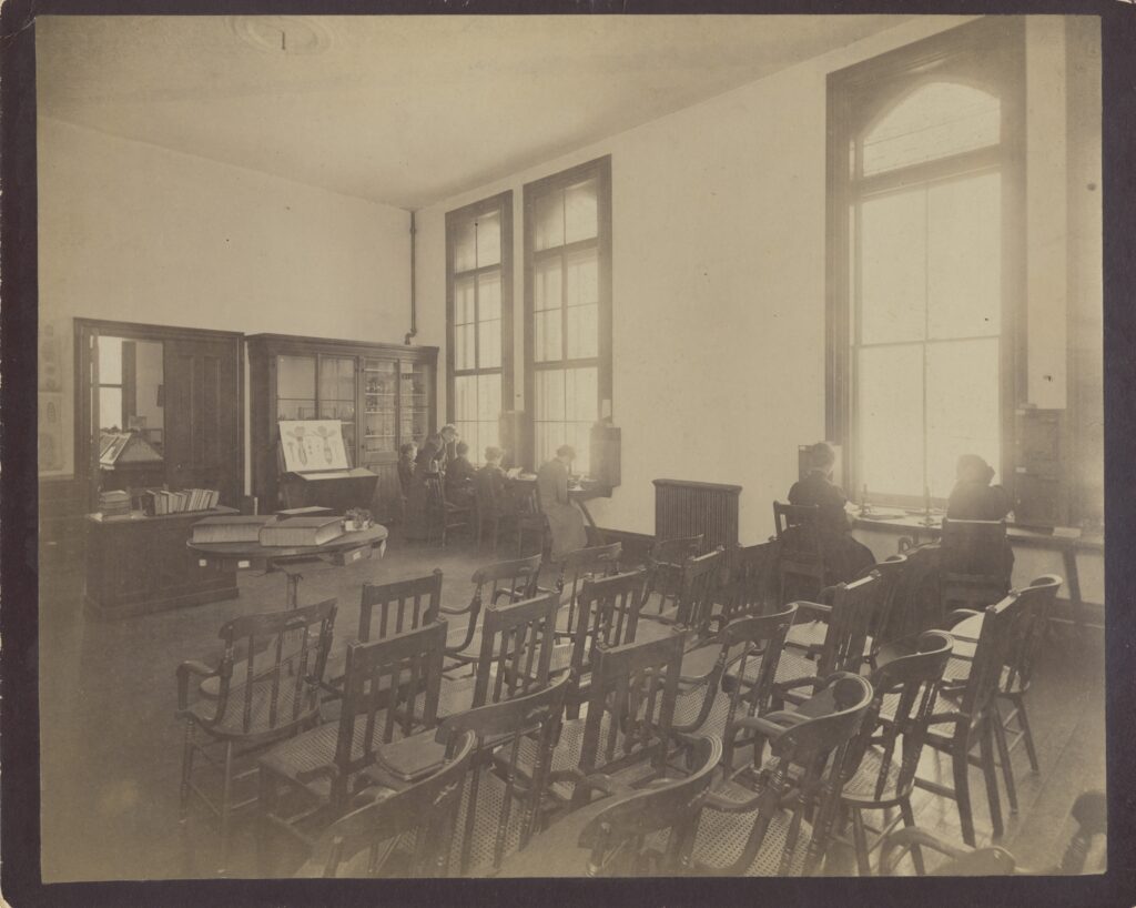 Black and white photograph of classroom with light coming in large windows. Rows of chair sit in the foreground, and students sit at tables set below the windows bent over microscopes.