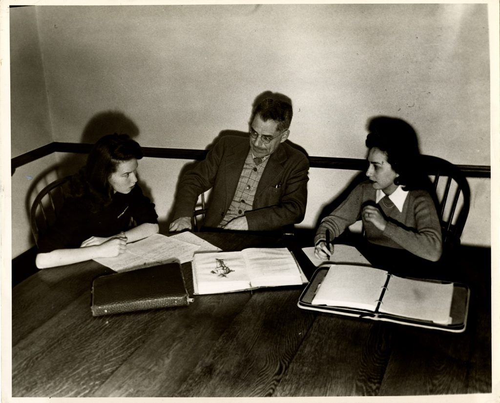 Two French students and a professor are immersed in conversation around a wooden table with binders and books in front of them.