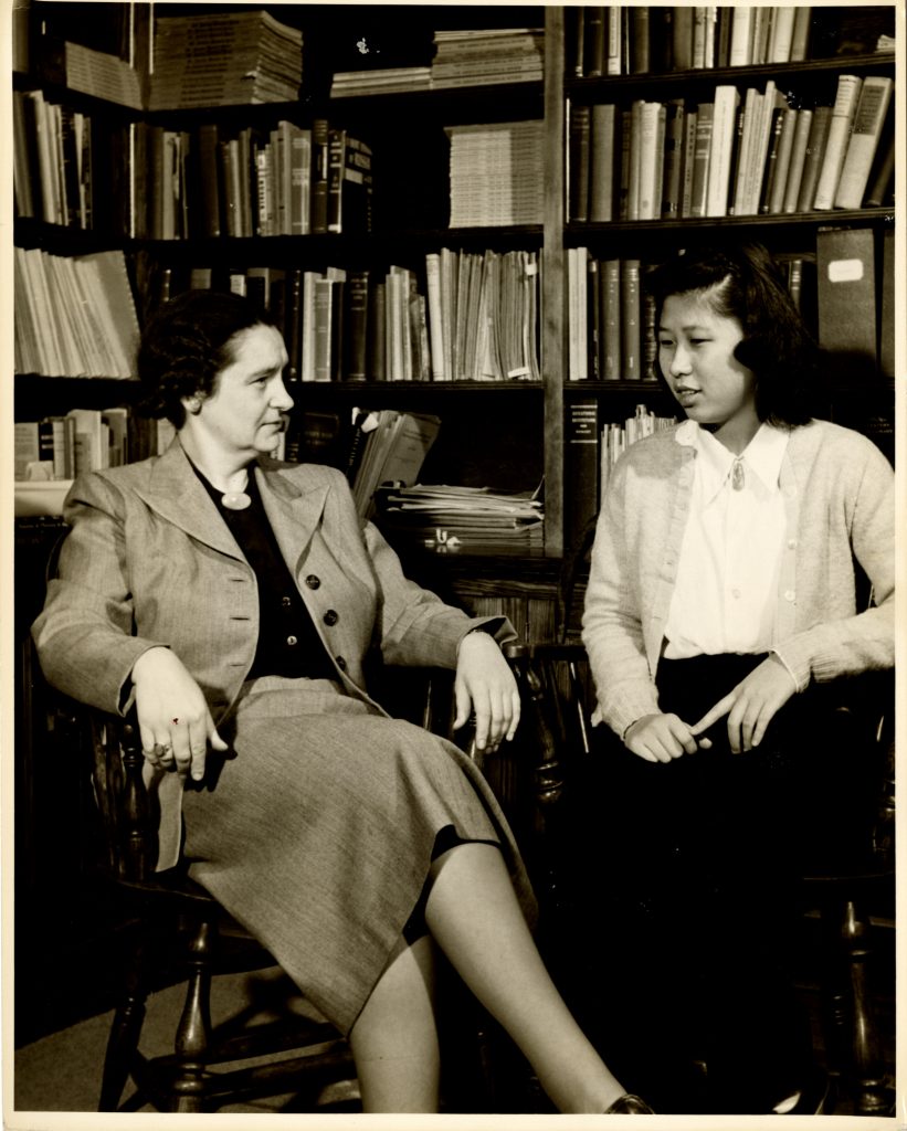 Moy Yoke Chong, former member of the class of 1953, wears a light-colored cardigan over a white blouse as she speaks with Academic Dean Meribeth Cameron in the an office, before a shelf of books.