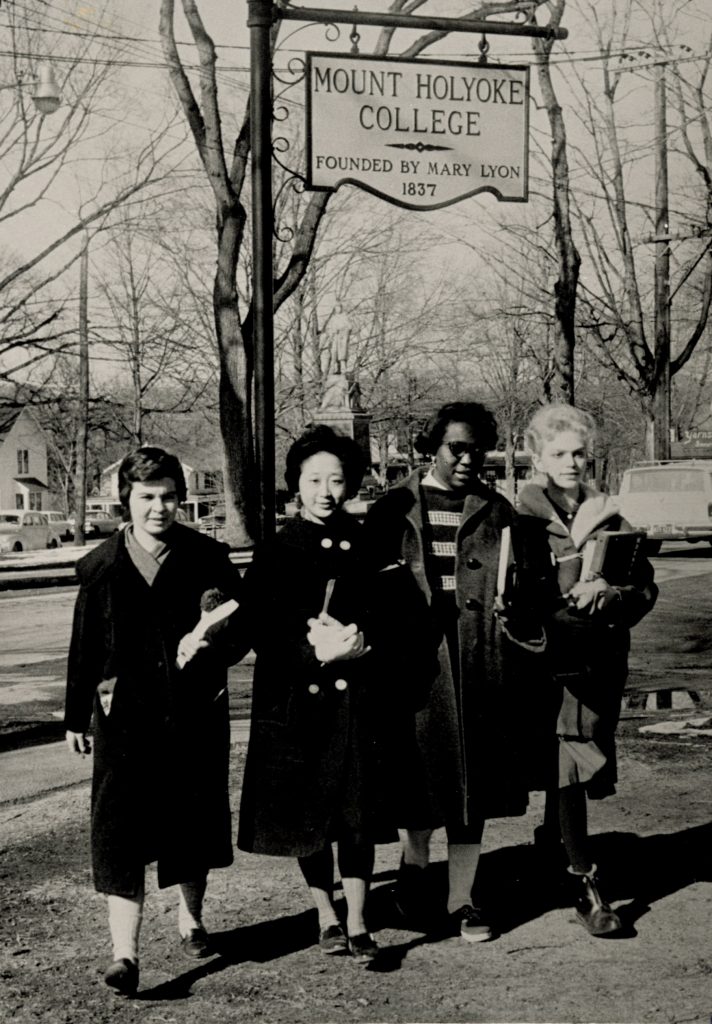 Four students in winter jackets with books walking outdoors beneath a Mount Holyoke College sign.