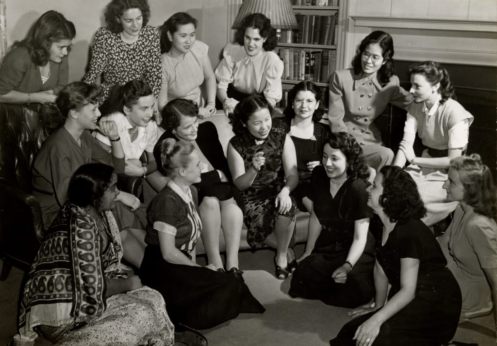 Sixteen students lounge and chat in a dormitory common room. Some gather around a sofa. Others are seated on the floor.