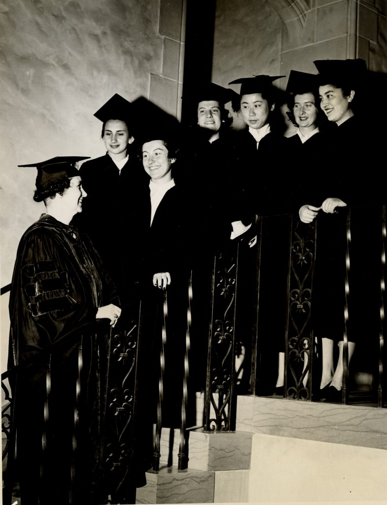 Six students in black graduation gowns and caps standing on a staircase with the academic dean.