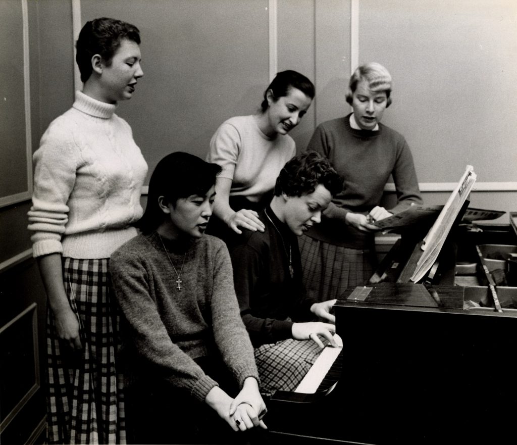 Five students gathered around a piano. One student plays the keys while the rest sing.