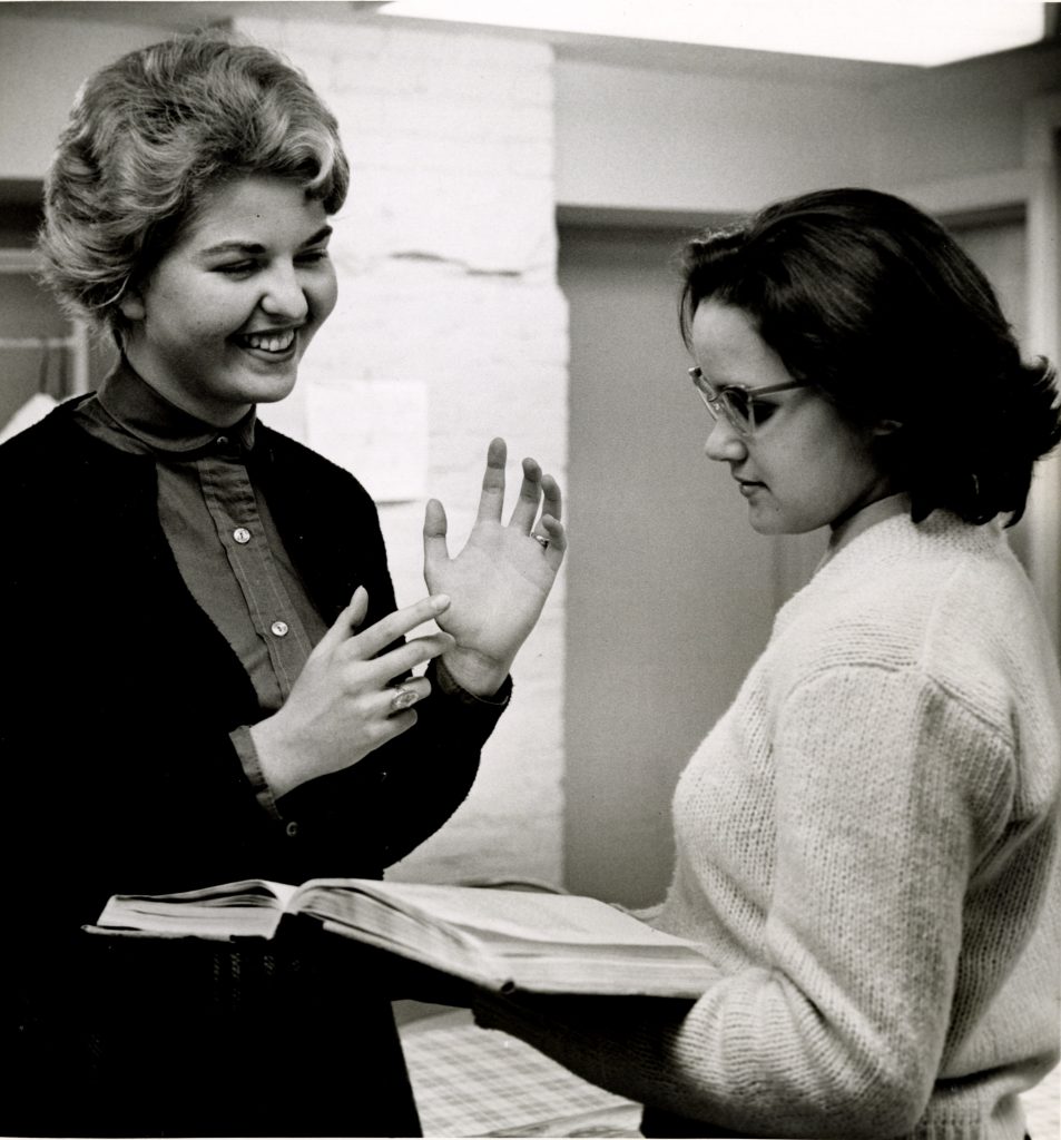 A student tutor smiling and gesturing as she explains something to her tutee, who holds a book.