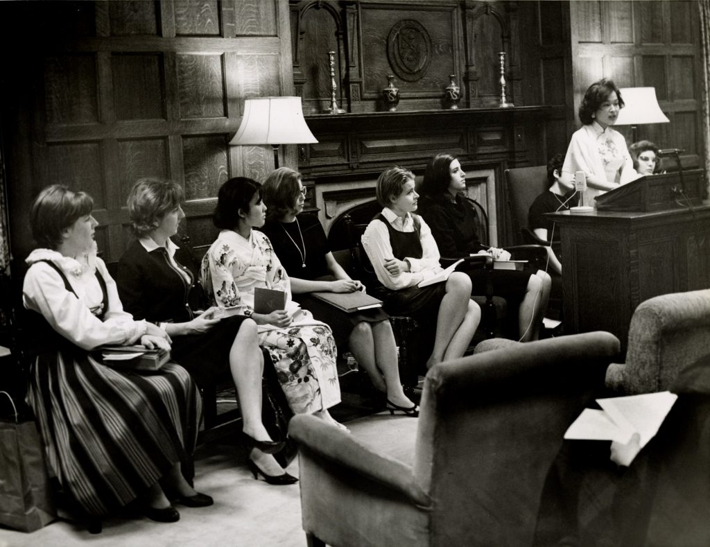 Nine students in formal attire seated in a row at the front of a poetry-reading event. One student stands at a podium to read.