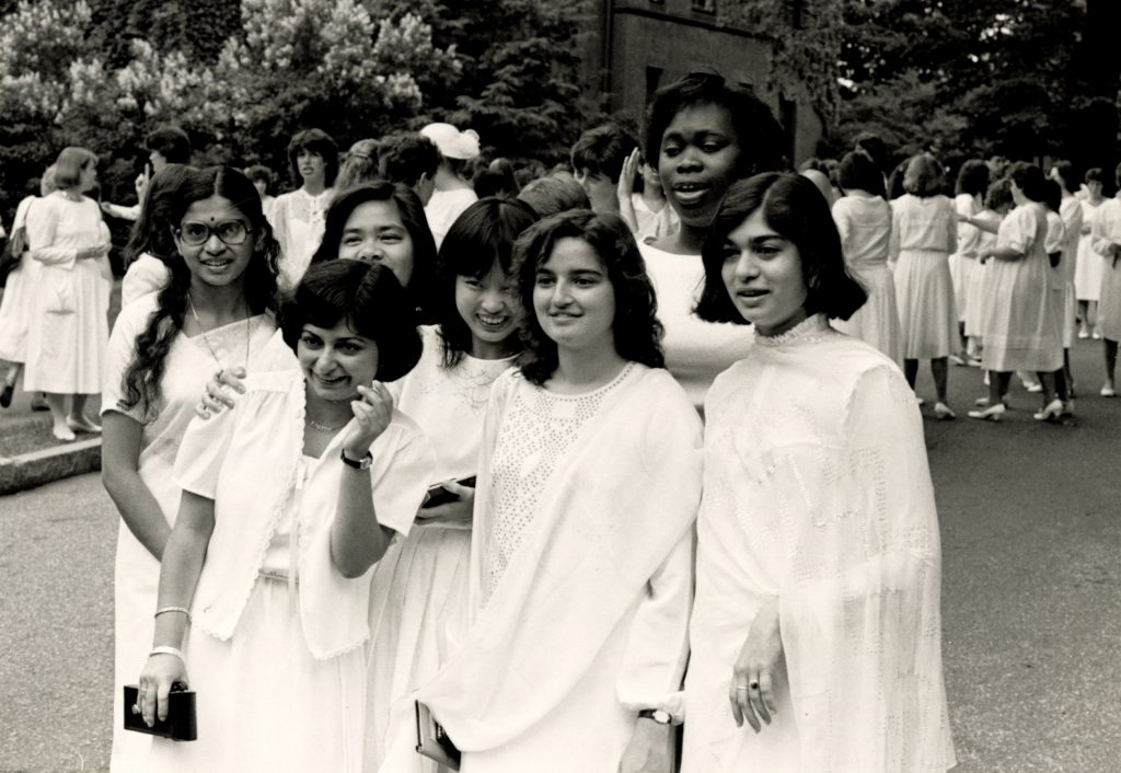 Seven students in white dresses smiling for a photo at the Laurel Parade. Similarly dressed students mingle in the background on the street.
