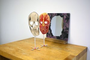 Two stands are lined up in front of a mirror.  On the first stand is a 2D skull made out of handmade kozo paper.  On the second is the muscles of the human face made of string and hand dyed kozo paper. Both have the eyes cut out so the viewer can look through into the mirror.