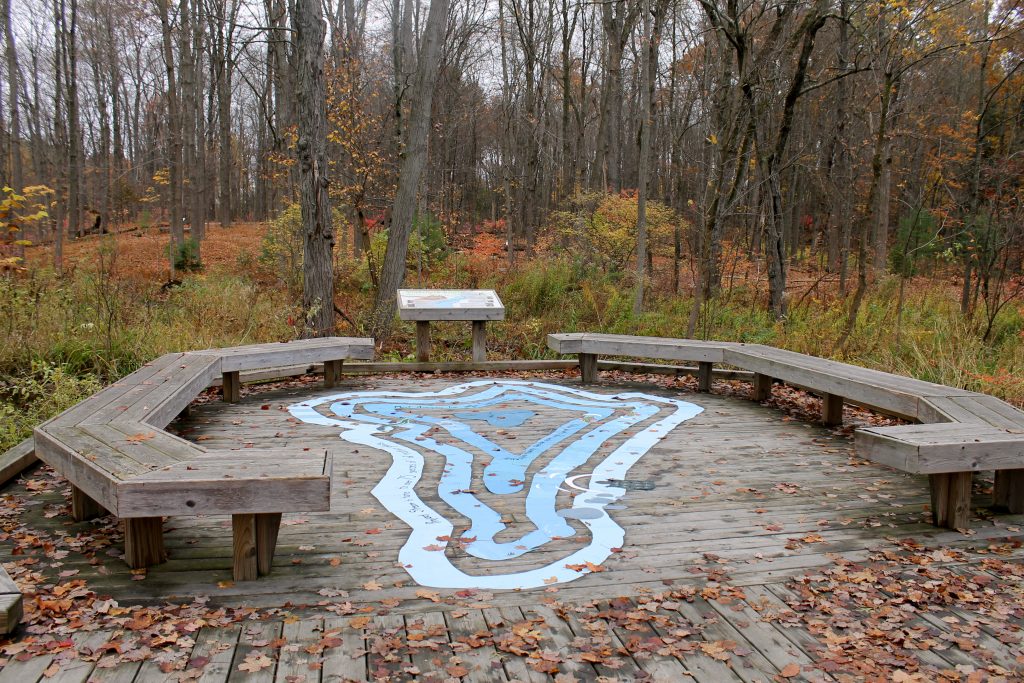 a print of an abstract lake with text lays on the ground in the center of a circle of benches, surrounding are trees and a stream.