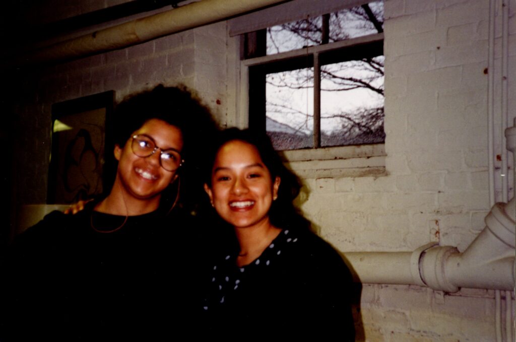 A color photograph of two students smiling for the camera with arms around one another. The background show a high, small window and pipes, showing that they are in a basement.