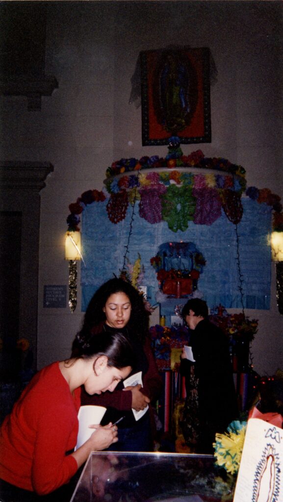Two students holding pens and paper looking down. In the background are colorful decorations for a Dia de Los Muertos event
