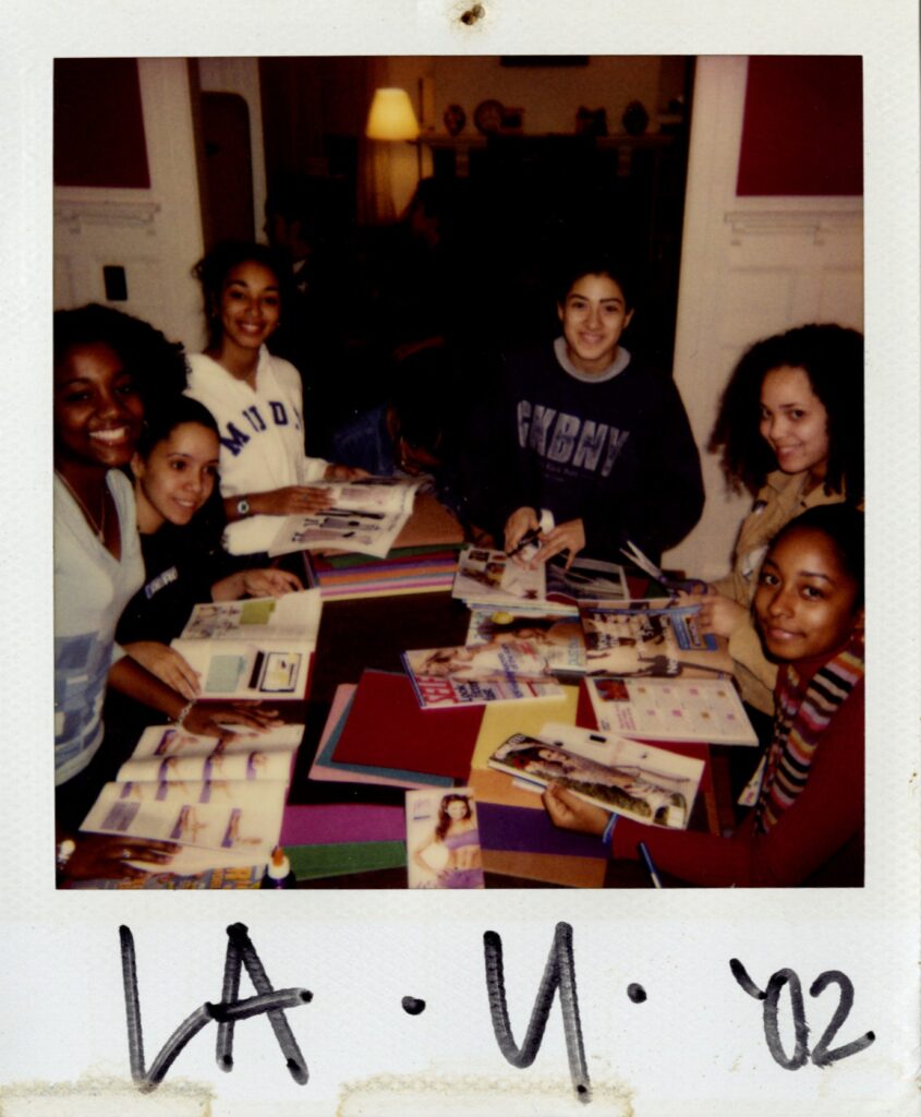Six students gather around a table and are smiling for the camera. The table is covered in magazines, construction paper, and scissors.