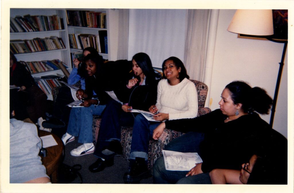 Photograph of six students sitting together on a couch and chair talking. One smiles for the camera. A full bookshelf is in the background.