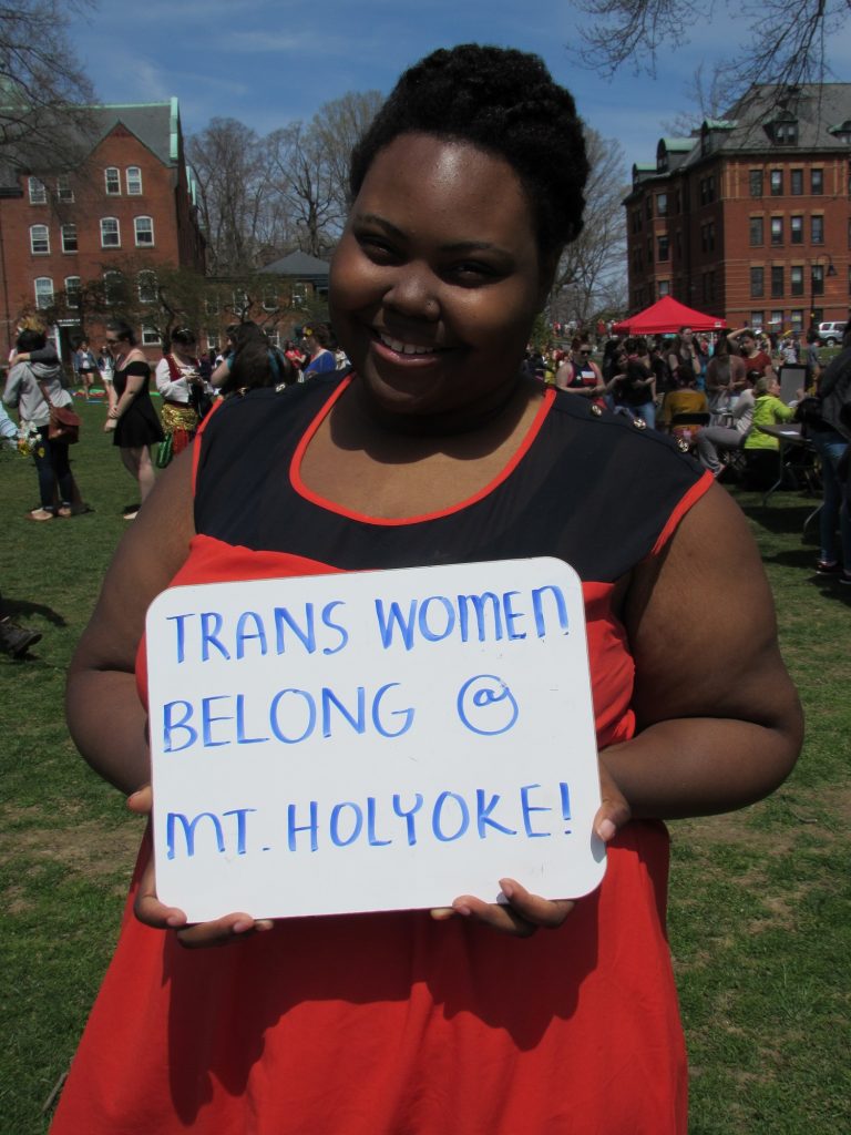 A Mount Holyoke College student stands on Skinner green holding a sign reading "Trans women belong @ Mt. Holyoke!"