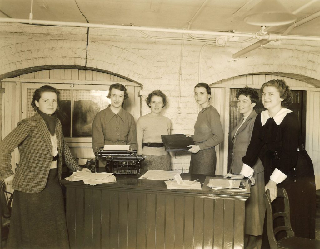 Six Mount Holyoke students standing around a desk. One of the students in the middle holds a leather briefcase out towards another student.