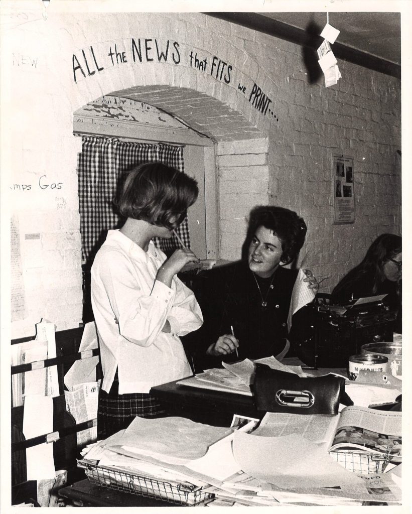 Two students at a desk. Painted on the brick wall behind the students are the words "ALL the NEWS that FITS we PRINT...."