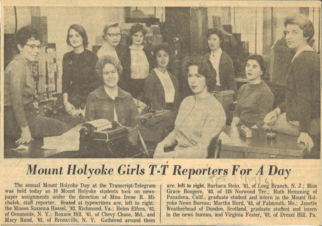 A group of students from Mount Holyoke sit on and at desks in the news office of the Holyoke Transcript-Telegram newspaper.