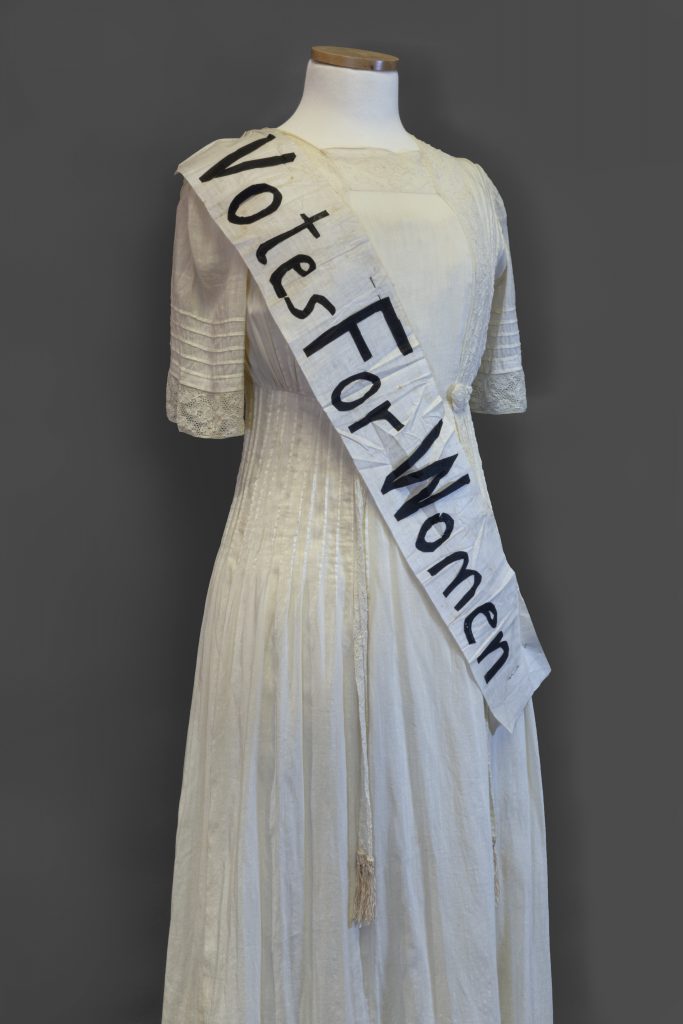 A mannequin angled slightly to the right wears an ecru cotton dress and a “Votes For Women” sash. The dress has a high square neck and elbow-length sleeves, both trimmed with lace. This lace continues vertically down the bodice with pintucking along the sides and narrows at a high waist with two lace rosettes. The lace is continued down the skirt with two decorative strips attached at the waist; they end above the knee in decorative tassels. The white sash is pinned diagonally at the right shoulder and left hip with black paper letters spelling “Votes For Women.” The letters are hand-cut and obviously imperfect. The sash is slightly creased between “For” and “Women” and the paper ‘W’ is beginning to pull up from the sash in some places where it was folded.