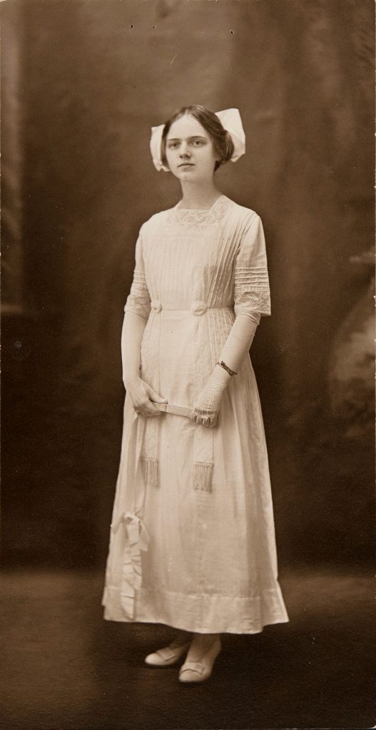 Louise Dunbar wearing the dress circa. 1912. Her hair is loosely pulled back and seems to be tied with a very large white bow. In addition to the dress, she wears a pair of white slippers, white stockings, elbow-length white gloves, and a bracelet on her left wrist. In her gloved hands she holds either end of a closed hand fan below her waist. A long satin ribbon trails from the end of the fan to the ankle-length hem of her dress. The dress is evidently the same as the one on the mannequin, albeit in better condition.