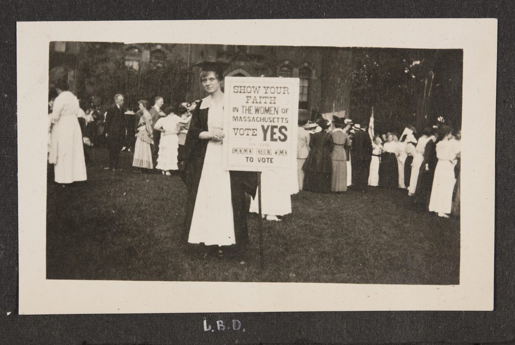 Louise Dunbar stands alone before a crowd of people wearing her black cap and gown over a white dress. She supports her "VOTE YES" sign with her right hand and grips it with her left fingers, looking meekly from the left corner. An unidentified brick building stands behind her. Other members of the mingling crowd appear to hold banners of unclear purpose as well.