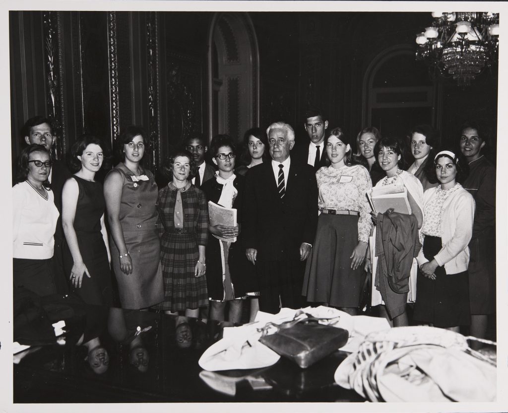 A group of 15 well-dressed students, mostly women, stand in a semi-circle in the midst of an ornate room. Senator Bill Keating stands in the center in a full suit. In the foreground of the photo various coats and a purse are visible on a glossy table.