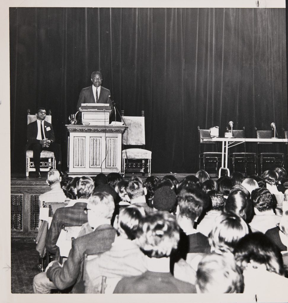 A man in a suit stands at an ornate podium on the stage of Chapin Auditorium. He smiles down at the crowd of students in their rows of chairs. Behind him to the left sits another suit-clad man in a high-backed chair. He wears glasses and clasps his hands in his lap, looking into the distance. To the right of the podium sits another high-backed chair. A folding table with microphones and a pitcher of water sits ready for panelists.