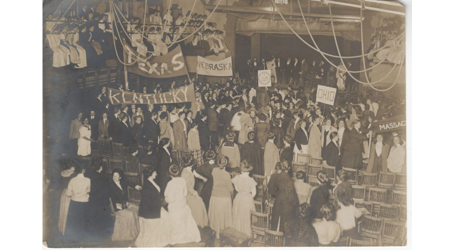 Hundreds of students crowd an early Blanchard Hall. Most stand on the ground floor among rows of wooden chairs, but many others watch from the balcony on the upper floor; those in the first row sit at the floor’s edge, clutching the railing as their feet dangle. The photographer likely stood on the balcony as well to obtain the view of the action. Thirteen other students, some dressed as male candidates, stand upon the stage at the background of the photo. Students within the crowd hold banners for Kentucky, Texas, Nebraska, and Massachusetts, and posters for Ohio and New York (represented by the state seal). At the top of the photo, gymnasium ropes, twisted out of the way, are visible.