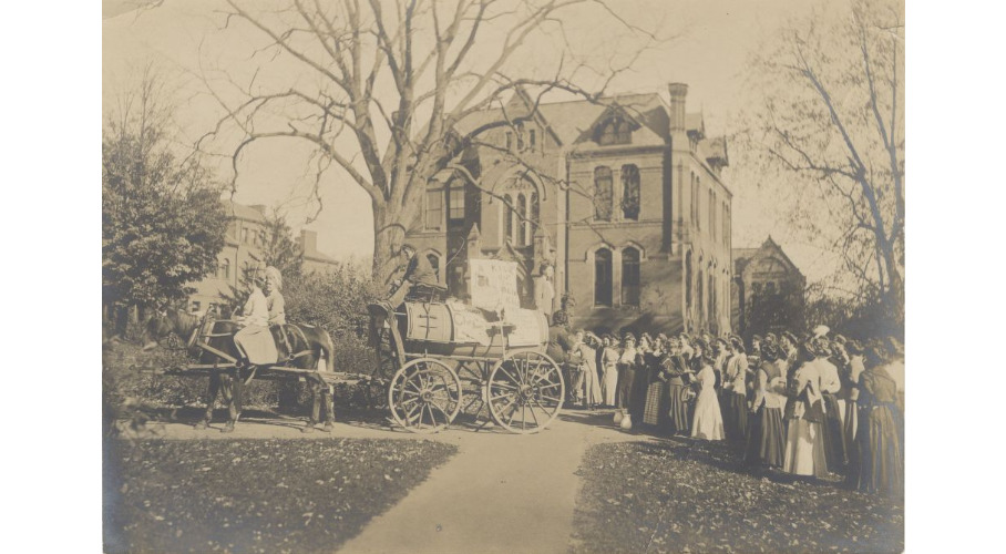 Two students facing left sit astride the horses harnessed to a wooden wagon carrying a large wooden barrel. Other students sit at the carriage’s rear, one standing to speak to the students clustered around her. Unfamiliar brick buildings are visible in the background, likely academic halls that have since been torn down. From the variety in foliage and the leaves littered on the grass, the photo was likely taken in the fall.