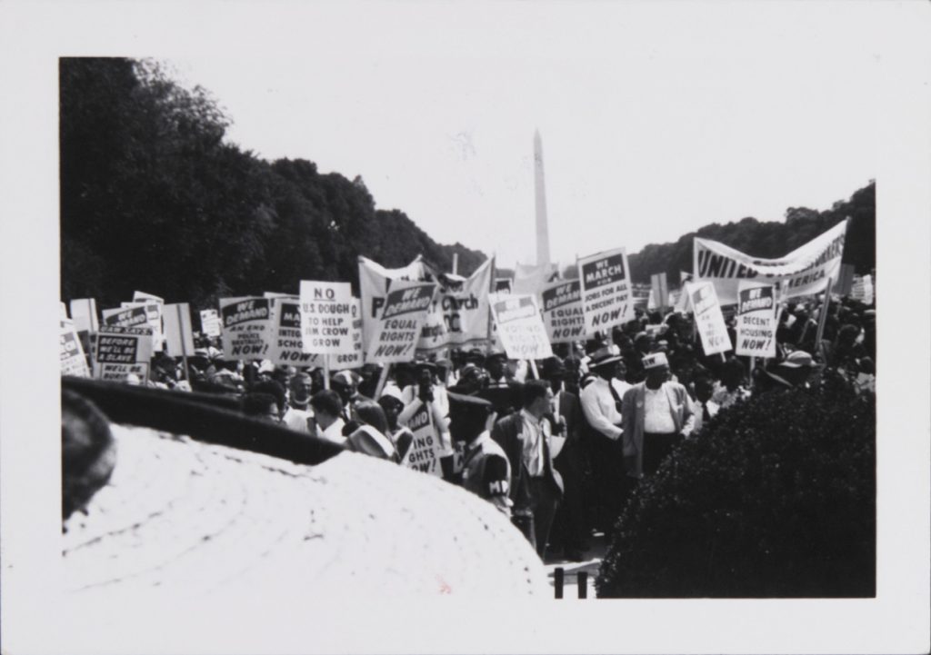 A predominantly Black crowd of adults stand on the National Mall. Many hold posters high on yardsticks, or long banners stretched between two poles with messages demanding racial equality. A sampling of the signs include: “WE DEMAND DECENT HOUSING NOW!” “WE DEMAND EQUAL RIGHTS NOW!” “WE DEMAND AN END TO POLICE BRUTALITY NOW!” “NO U.S. DOUGH TO HELP JIM CROW GROW.” What appears to be a wicker hat and a bush in the foreground obscure some of the crowd. The Washington Monument is visible in the background, rising between the lines of trees.