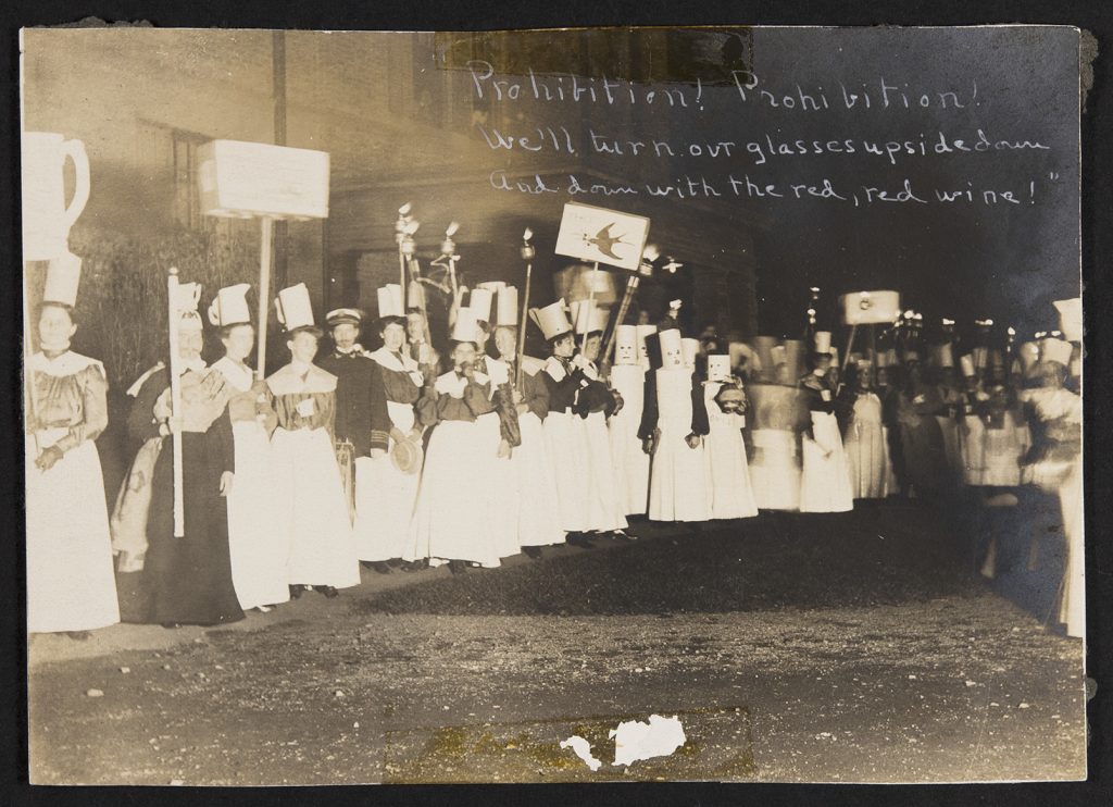 Many students variously attired and holding signs congregate outside a brick building; one student peers down at the parade from a window on the second story. Several of the students wear matching blouses, white skirts, and paper “pitchers” on their heads. Some carry torches aloft; others posters of pitchers or advocating for “peace.” Most are not readable due to overexposure. Some students are clad in all white with cylinders of paper worn as masks with cutouts for their eyes, nose, and mouth. Others wear military garb over their dresses and have mustaches and goatees drawn on their faces. The line of students continues on in a blurry and semi-indistinguishable fashion. In white ink in the photo’s upper right corner is written “‘Prohibition! Prohibition! / We’ll turn our glasses upside down / And down with the red, red wine!’”