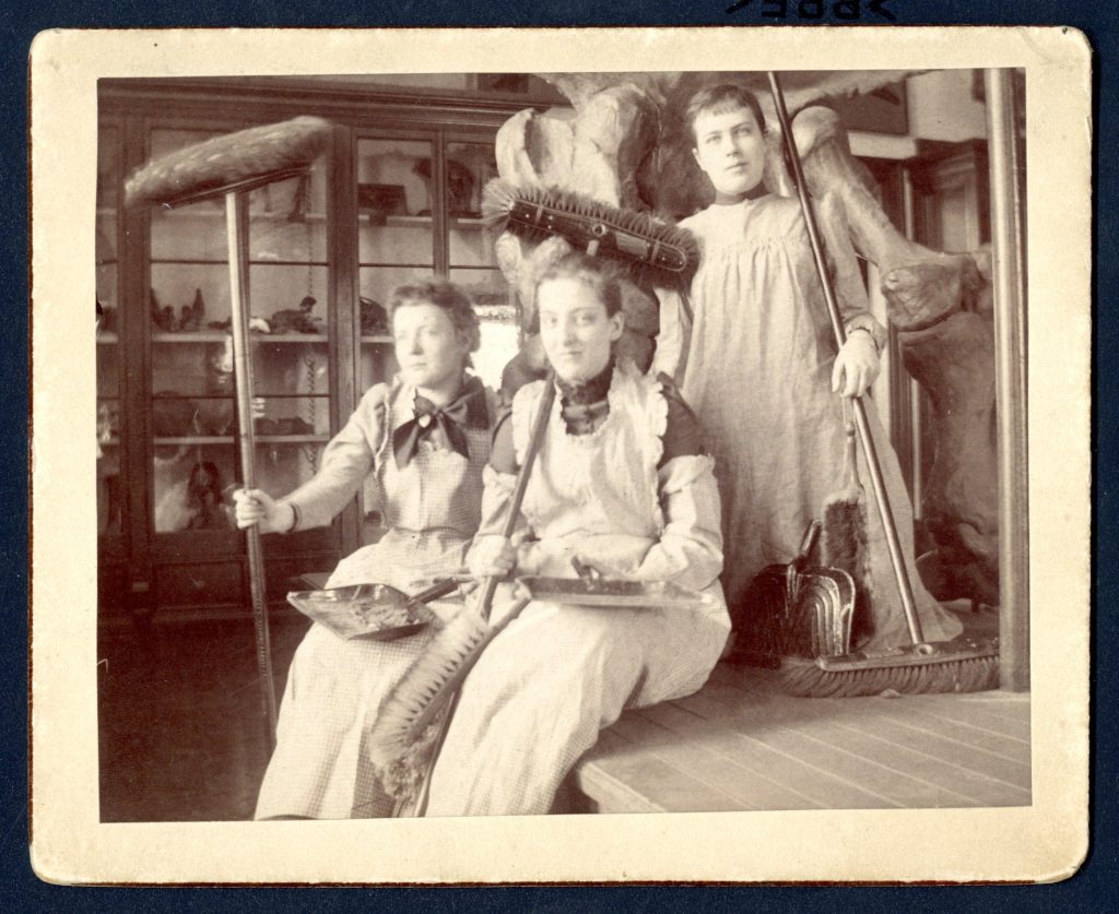 Two women seated, one on her knees, holding brooms and dust pans