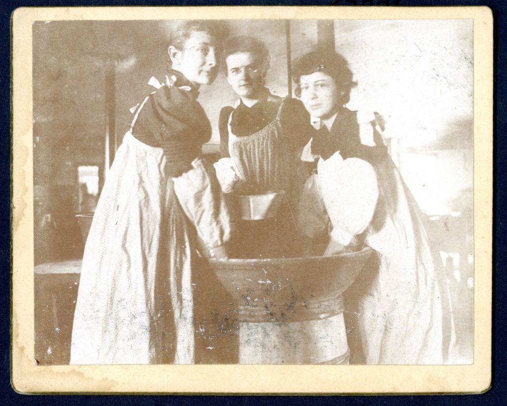 Three women stand around a large mixing bowl wearing aprons with their hands in the bowl.