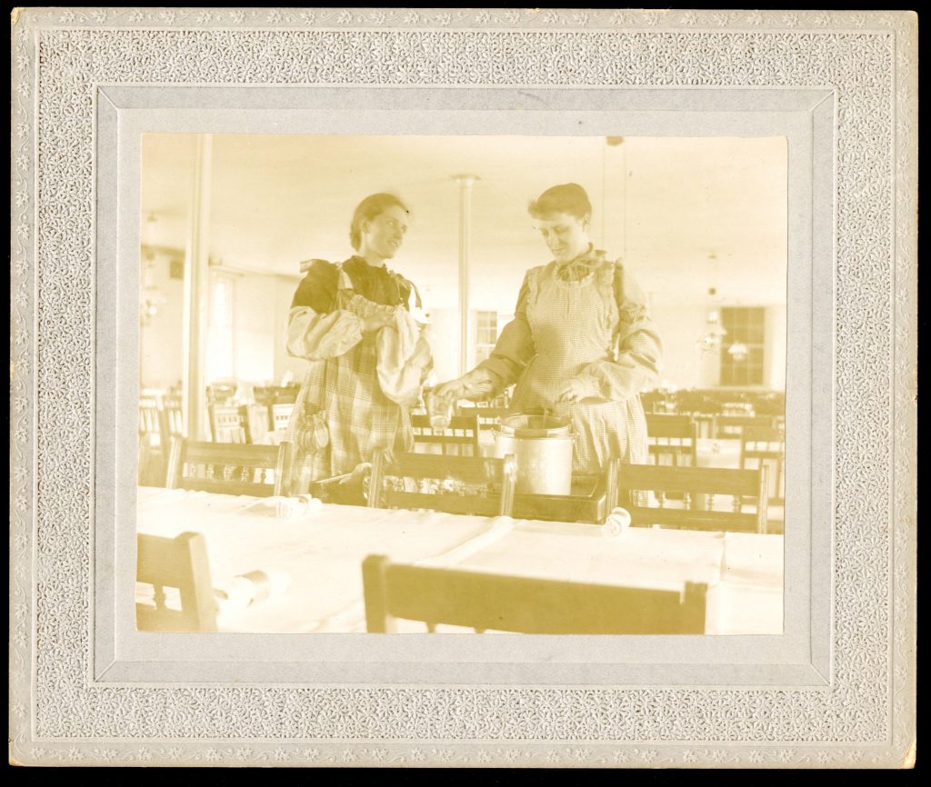 Yellowed black and white photograph of two students in aprons washing glasses on a cart between rows of long tables and chairs.