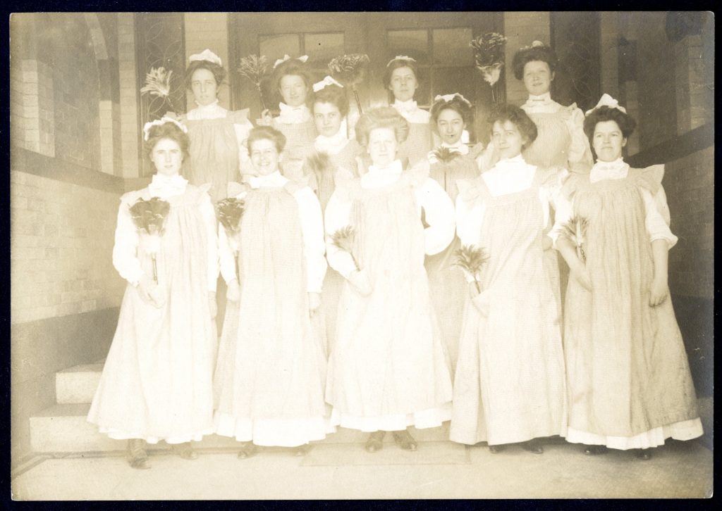 Students in cleaning uniforms holding feather dusters pose together on steps outside a building