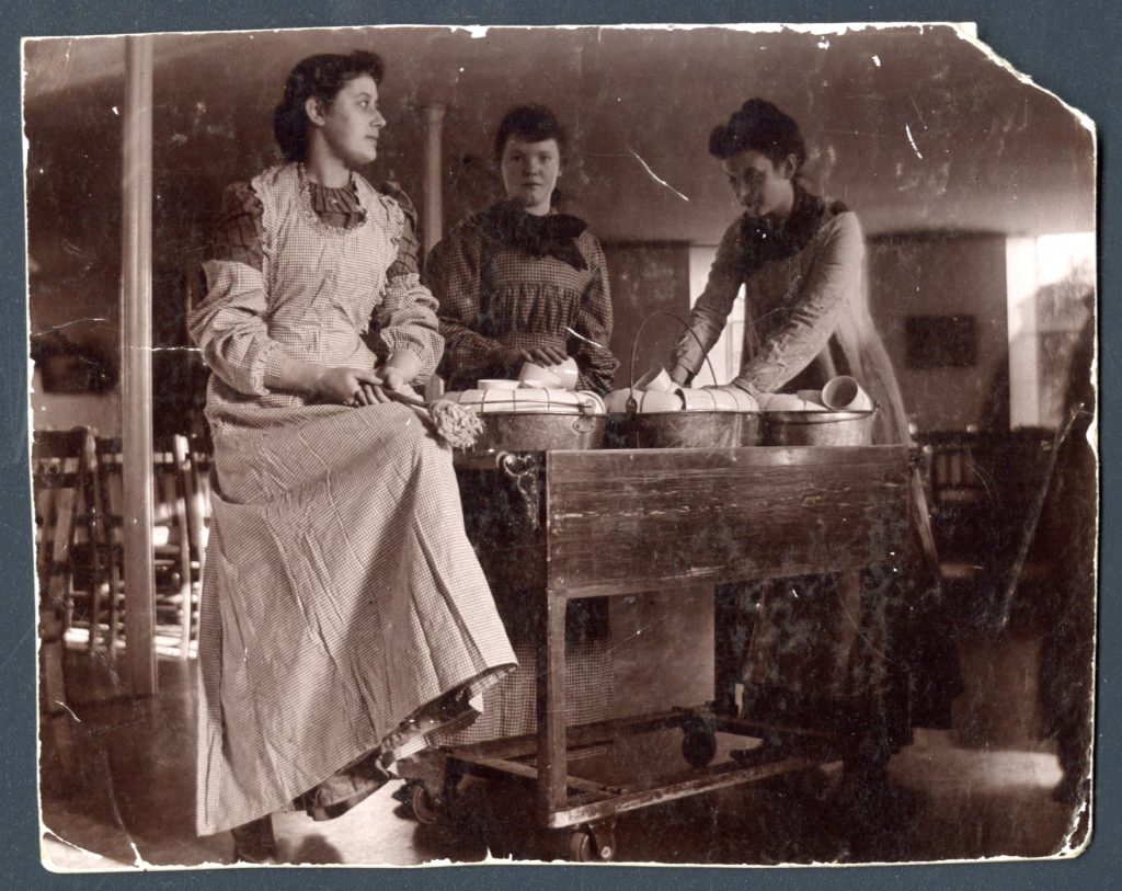 Three students stand around a table with bins filled with stacked dishes