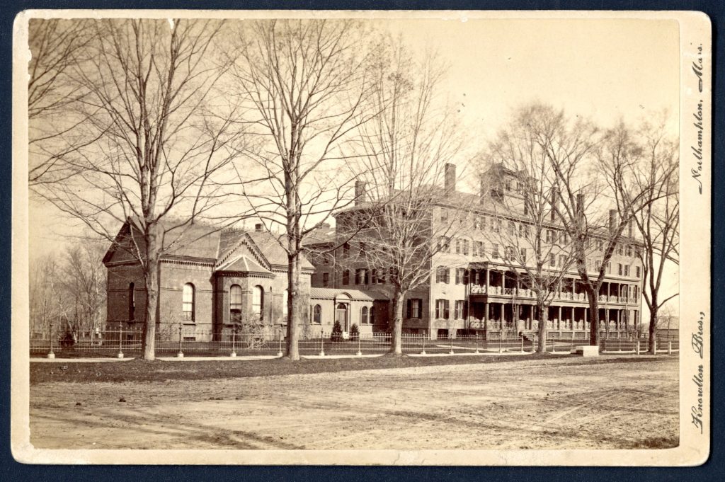 Street view of a large four story brick building with a two story outdoor porch. Connected on the left is a smaller, two story building.