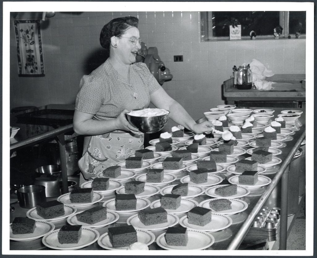 A woman in a hair net and apron spoons whipped cream on rows or sliced cake on plates.