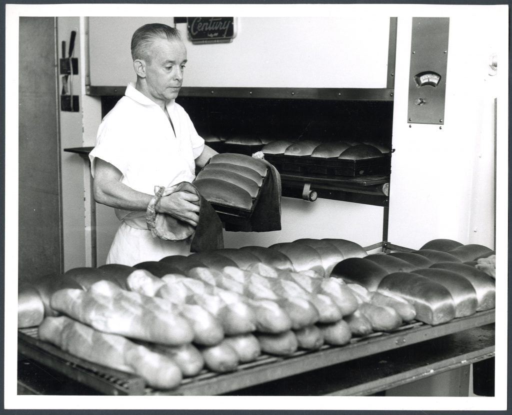 A man stands holding a tray of freshly baked bread. Behind him is the large oven, and in front of him is a table covered in bread.