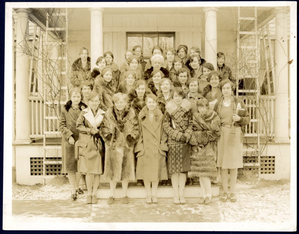 A group of students in jackets stand outside a house posing for the camera. Amidst them is an older woman, their housemother.