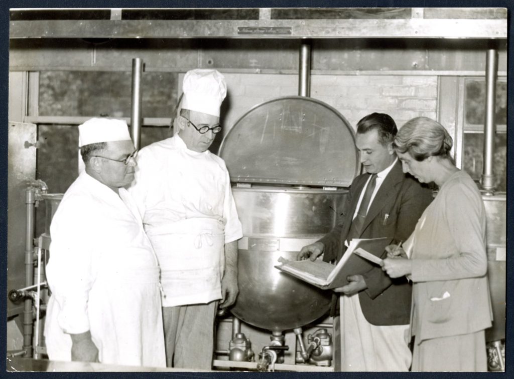 Four people, two in chef's uniforms, the other two in work attire, talk in front of a large cooking vat.