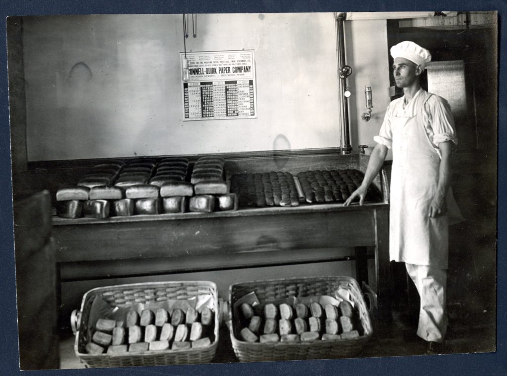 Black and white photograph of a man in an apron and chef's hat standing beside trays and baskets filled with bread.