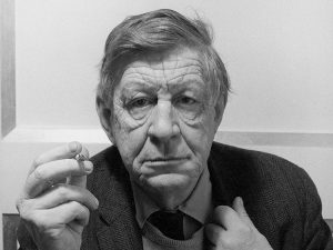 Portrait of American poet W. H. Auden (aka Wystan Hugh Auden) looking into the camera holding a cigarette. He is older in this photo, and is wearing a dark suit.