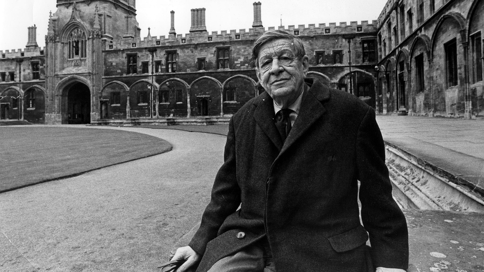 A black-and-white photo of W.H. Auden sitting in front of Christ Church College, Oxford.