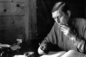 W.H. Auden sitting at his desk writing. He is smoking a cigarette with his other hand. The photo is in black and white.